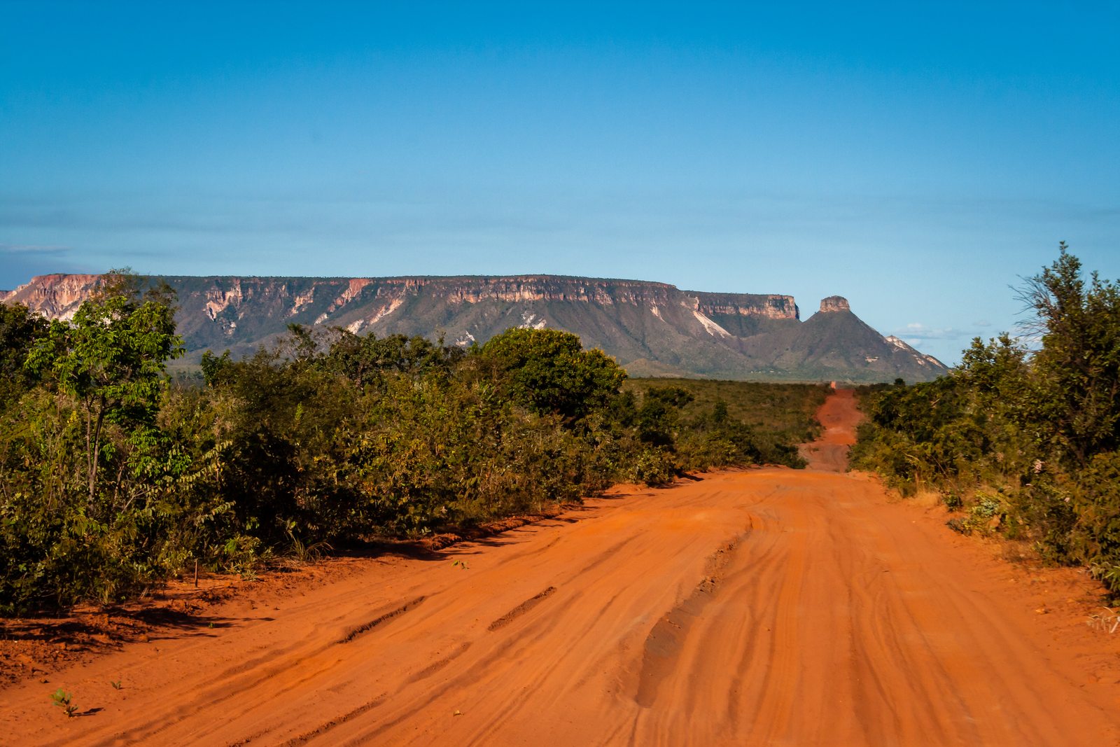 Estrada de terra avermelhada entre a vegetação do cerrado, com a Serra do Espírito Santo ao fundo, no Jalapão, Tocantins.